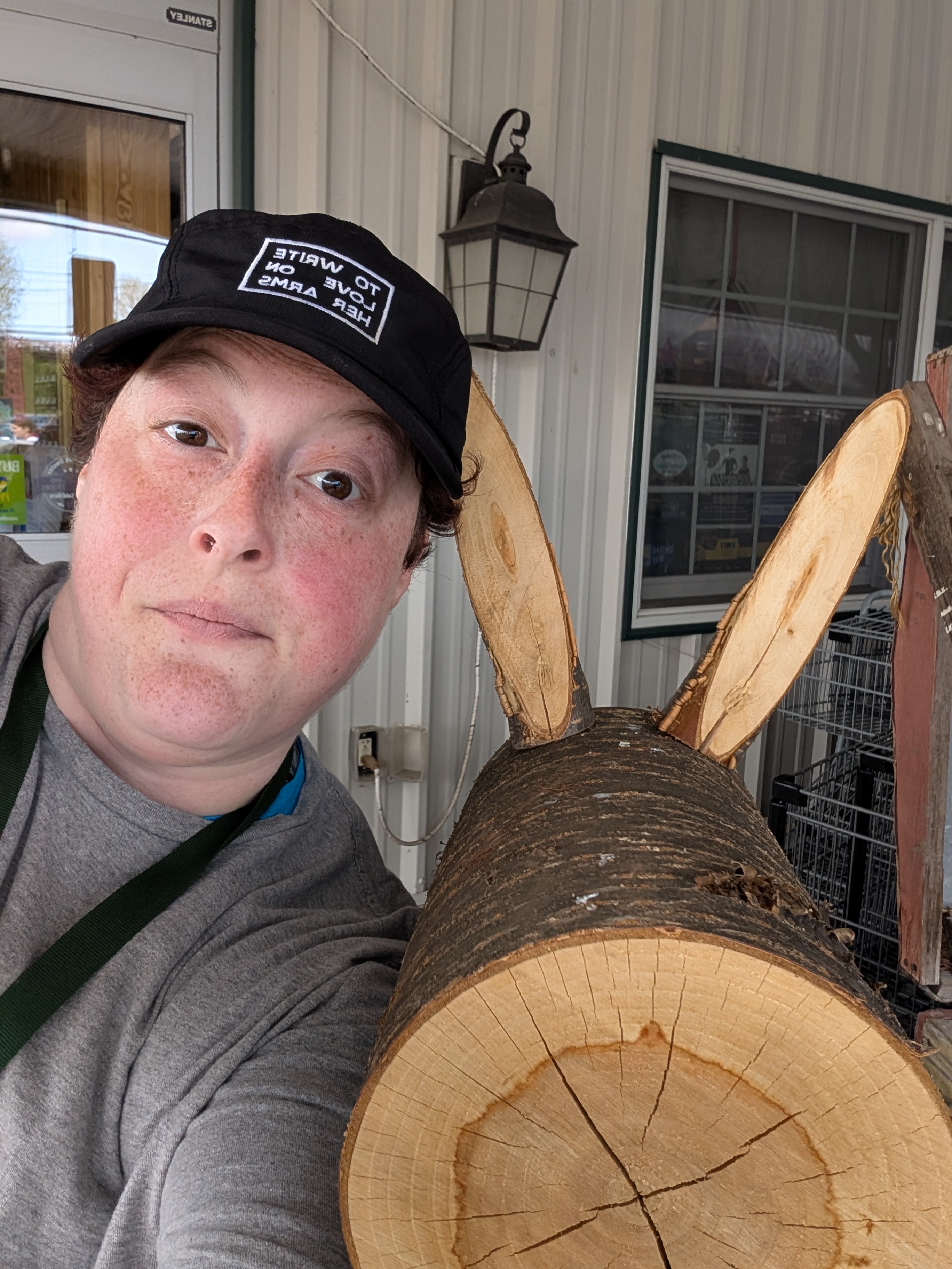 Nonbinary white person wearing a black hat faces the camera with a bemused expression. Next to them is a wooden reindeer.