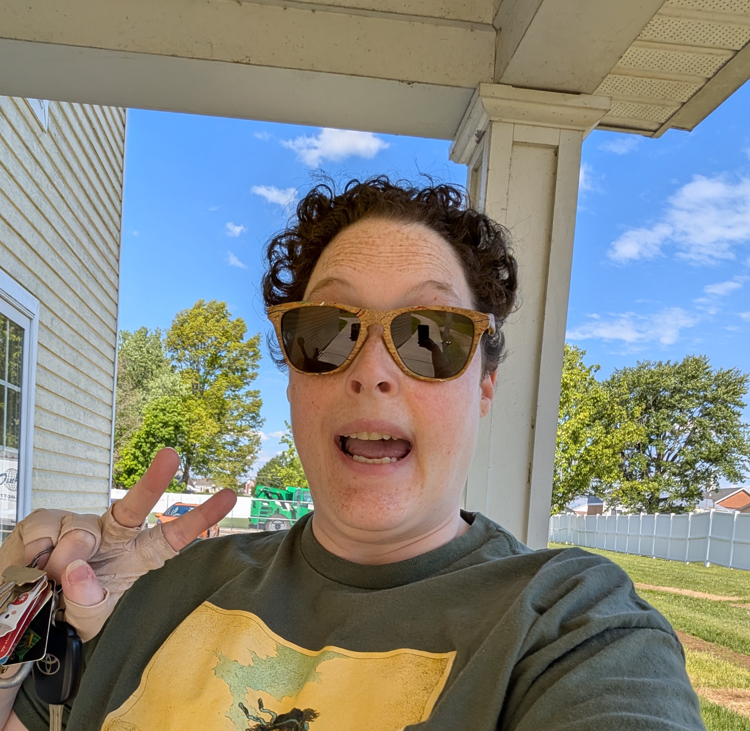 Nonbinary white person with short dark curly hair, wearing sunglasses, standing outside and making a 
